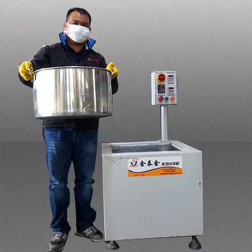 The image shows a man wearing a black jacket, blue jeans, and a face mask, holding a large stainless steel pot in his hands. He is standing in front of a machine with a white background.